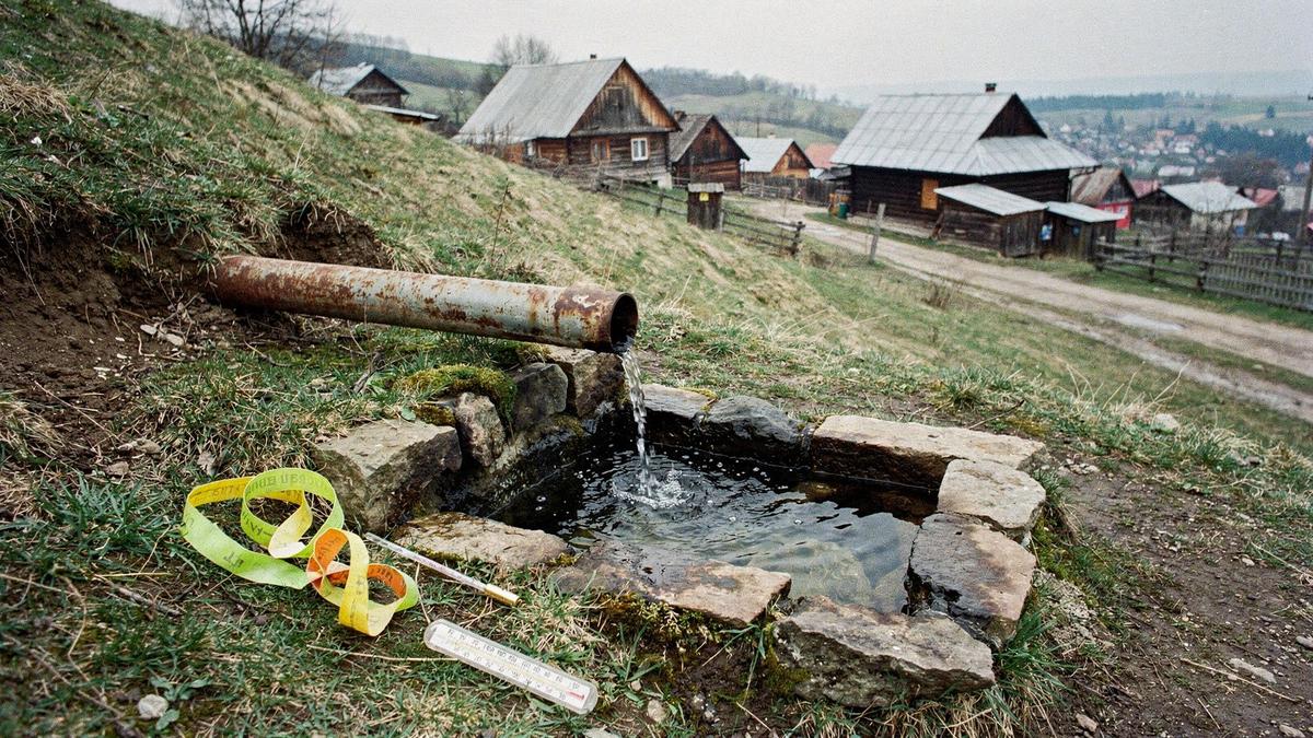 Mineral spring outlet piped into a stone basin above a Carpathian village, with a survey tape beside it